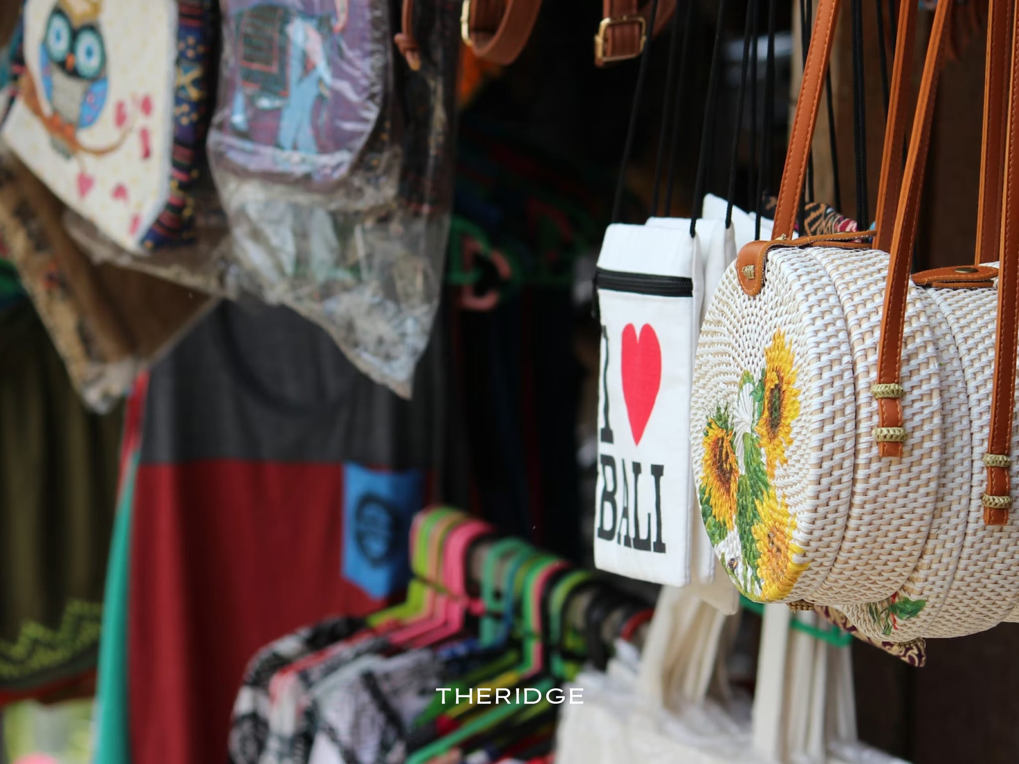 A close-up of traditional handmade souvenir bags, including a round woven rattan bag with a sunflower design and an "I Love Bali" tote, hanging at a stall in the Ubud Market.