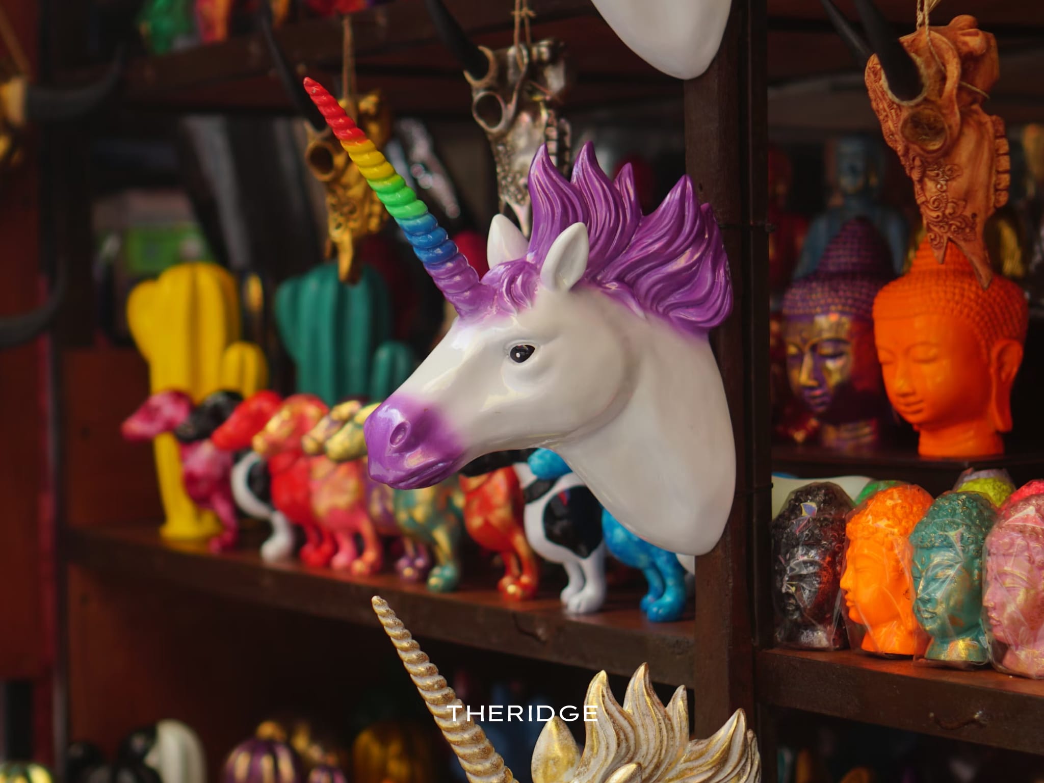 Colorful Balinese handicrafts featuring a purple unicorn head and vibrant Buddha statues displayed on a wooden shelf for sale at the Ubud Market.